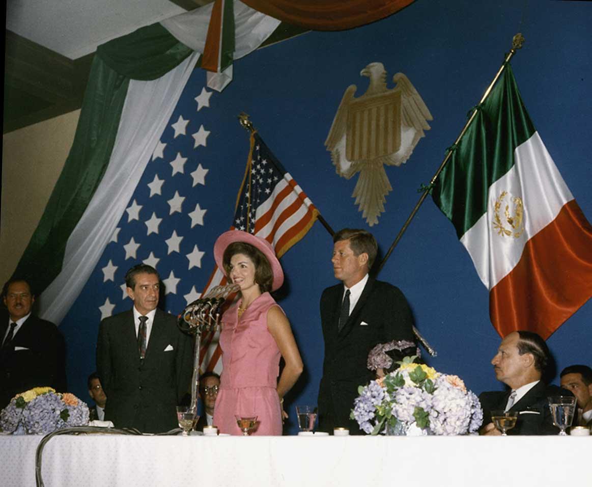 President Kennedy stands with First Lady Jacqueline Kennedy as she delivers remarks in Spanish at a luncheon during the presidential visit to Mexico, June 30, 1962