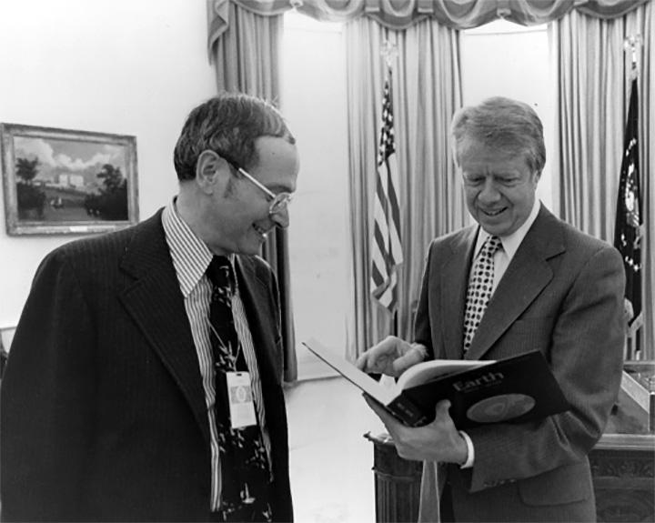 Frank Press looks on as President Jimmy Carter flips through Press's textbook.